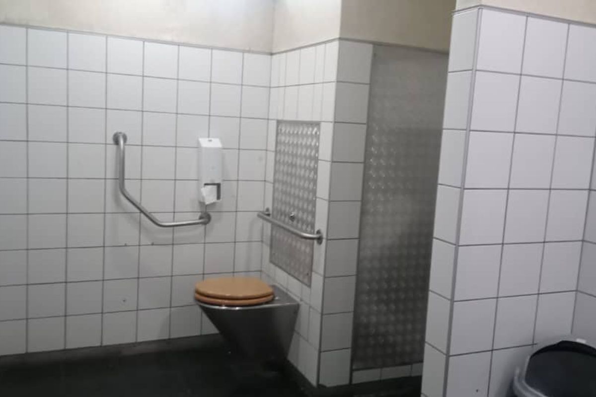 A functional office restroom featuring white wall tiles, stainless steel toilet fixture, and accessibility grab rails.