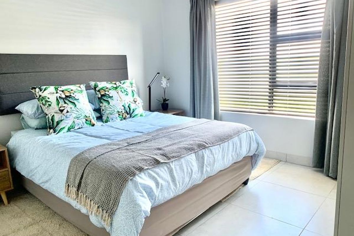 A bright and well-maintained bedroom featuring neutral tiled flooring, a large window with fitted blinds, and a modern upholstered headboard.