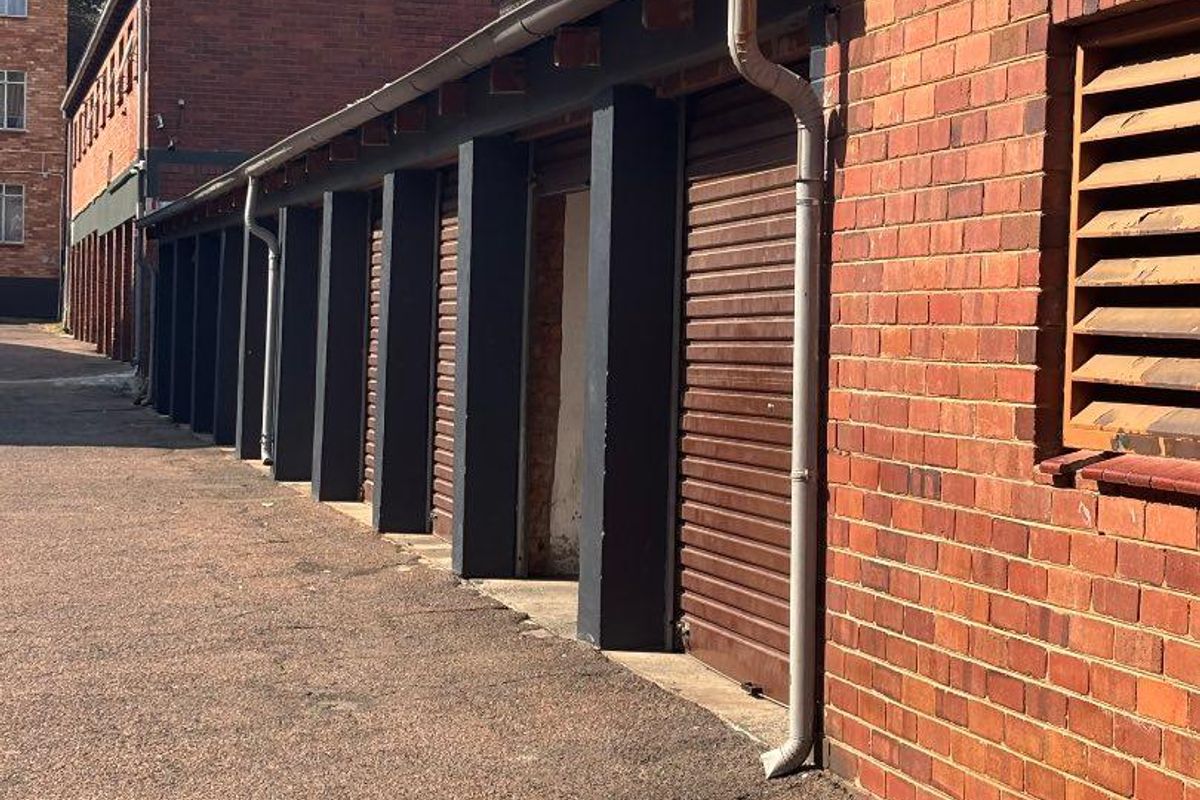A row of brick-built garages featuring roller shutter doors, external guttering, and downpipes, situated along an asphalt driveway.