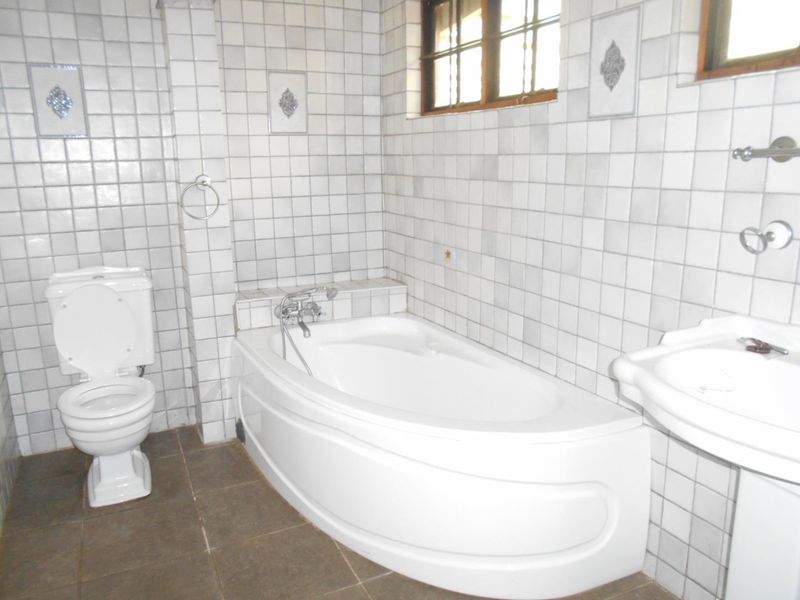 A dated bathroom featuring a built-in corner bathtub, pedestal basin, and toilet, finished with white wall tiles and dark floor tiles.