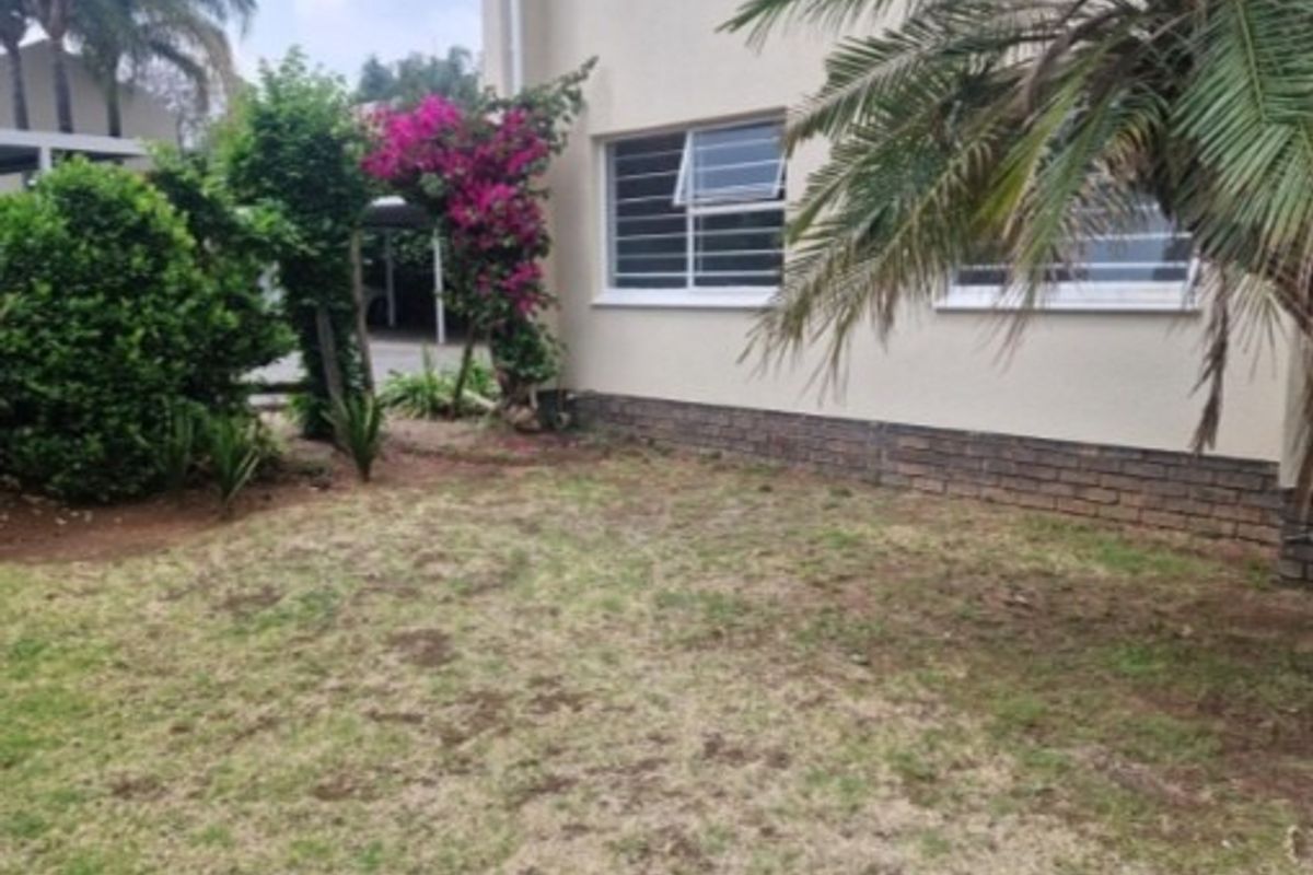 A view of the rear exterior of the apartment building featuring a small garden area, brick foundation detailing, and security-fitted windows.