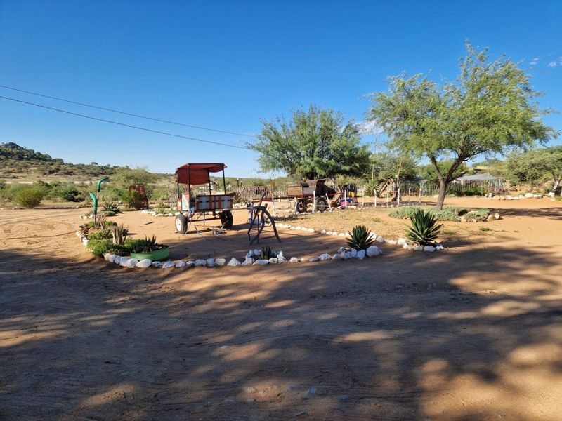 A spacious outdoor garden area featuring natural sandy terrain, mature trees, and landscaped garden beds bordered by decorative stones.