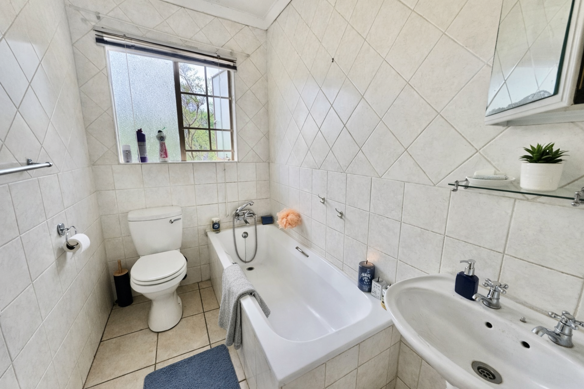 A functional bathroom featuring a standard bathtub, pedestal sink, and toilet with full-height wall tiling and a window for natural ventilation.