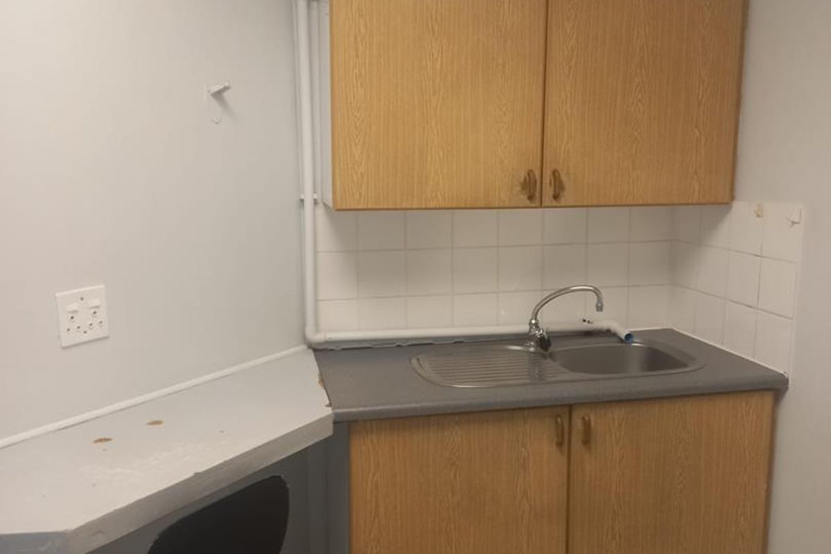 A compact office kitchenette featuring wood-look cabinetry, a stainless steel sink, tiled splashback, and a small attached counter space.