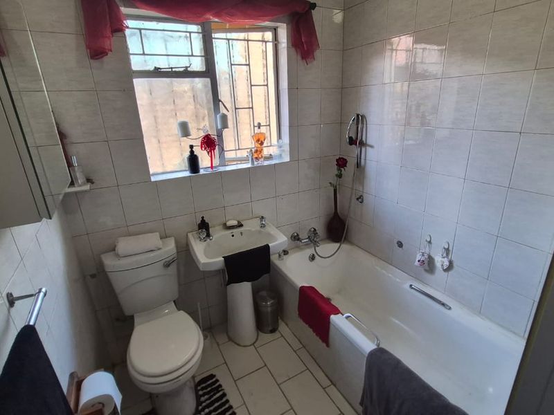 A functional bathroom featuring a pedestal basin, bathtub, and toilet with full-height wall tiling and a window with security bars.