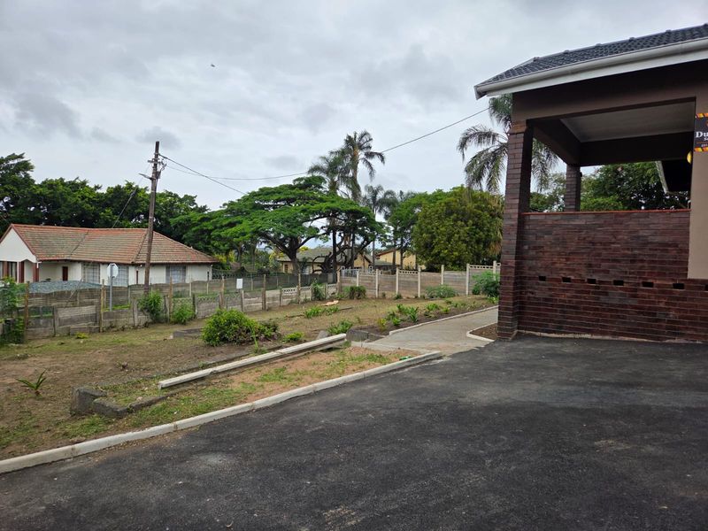 Rear exterior view of the property featuring a brick facade, concrete driveway, and a large, open garden space enclosed by a boundary wall.