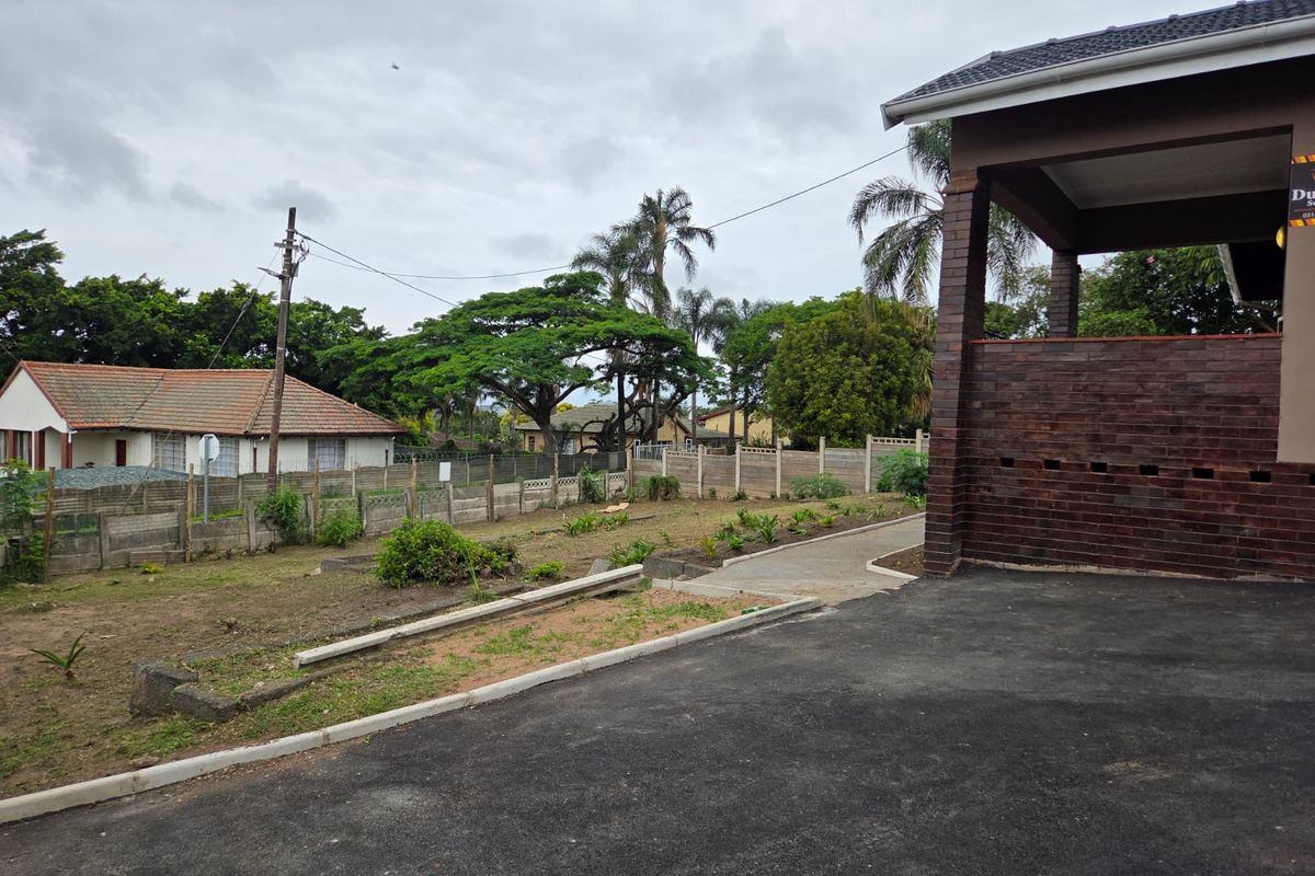 Rear exterior view of the property featuring a brick facade, concrete driveway, and a large, open garden space enclosed by a boundary wall.