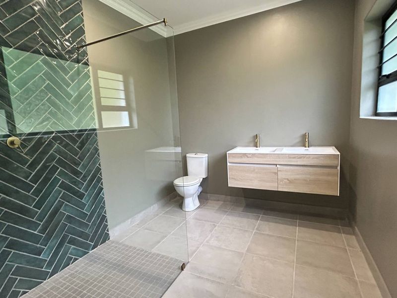 Modern bathroom featuring a walk-in shower with green herringbone tiles, a floating double vanity with gold fixtures, and neutral floor tiling.