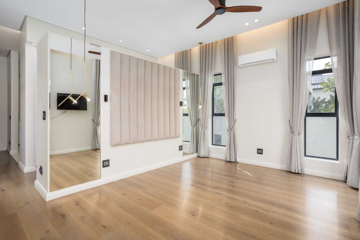 A modern, bright bedroom featuring engineered wood flooring, a ceiling fan, split-system air conditioning, and stylish mirrored and upholstered wall accents.