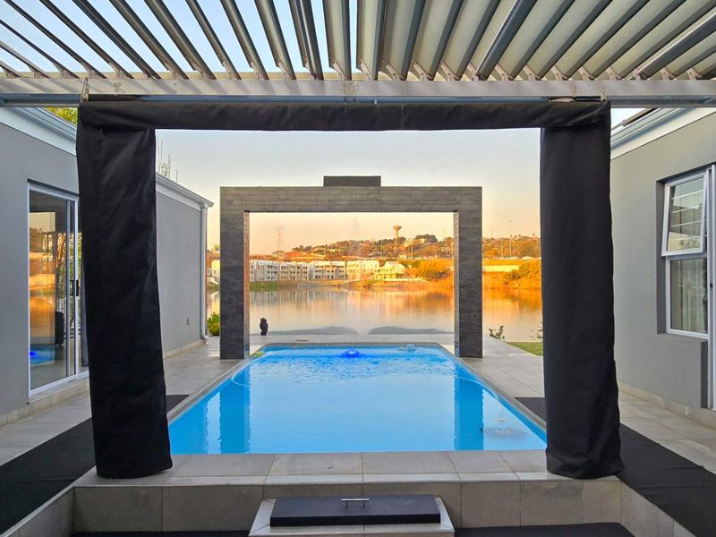 A modern outdoor pool area featuring a louvered pergola, stone archway, and direct access to the residence through sliding glass doors overlooking the water.