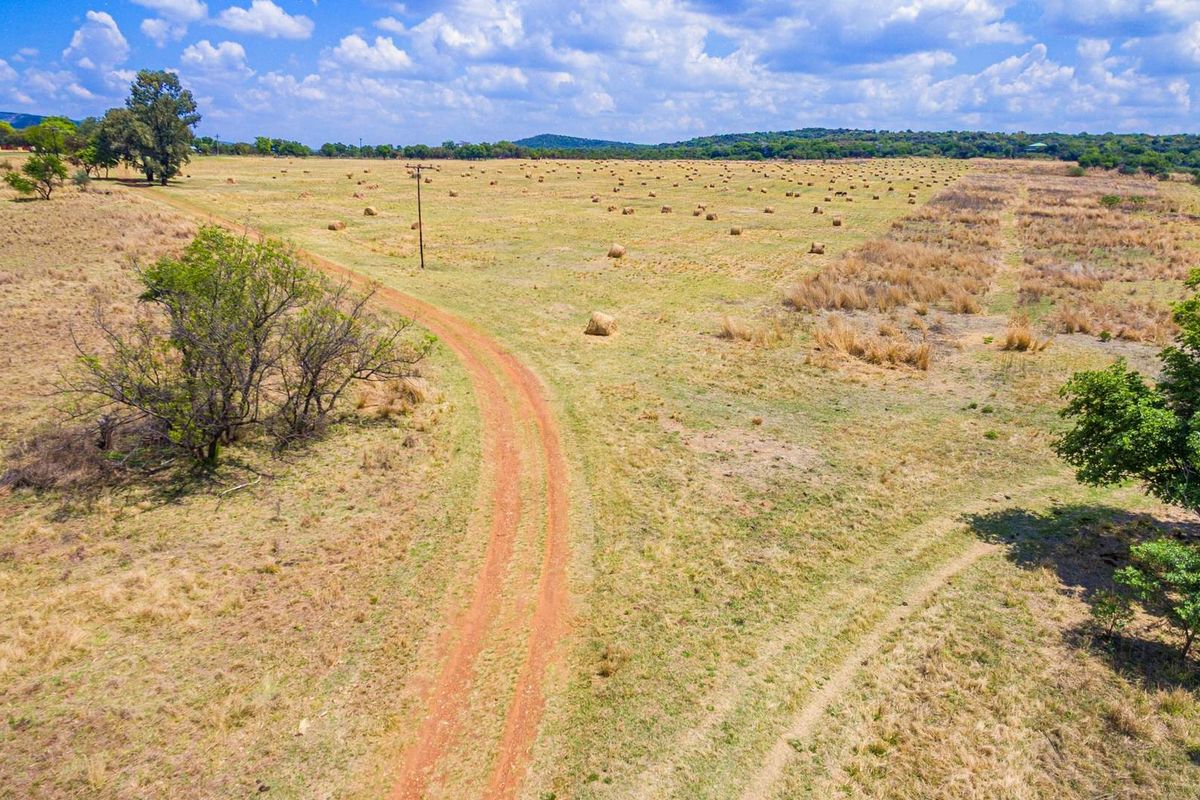 An aerial view of a large, open farm landscape featuring a dirt access road and scattered hay bales across the field.
