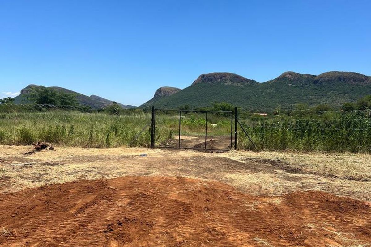 An expansive outdoor plot featuring a metal gate entrance, natural vegetation, and scenic mountain views in the background.
