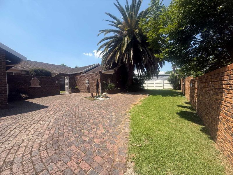 A front exterior view of the property featuring a brick-paved driveway, established garden, and brick boundary walls.