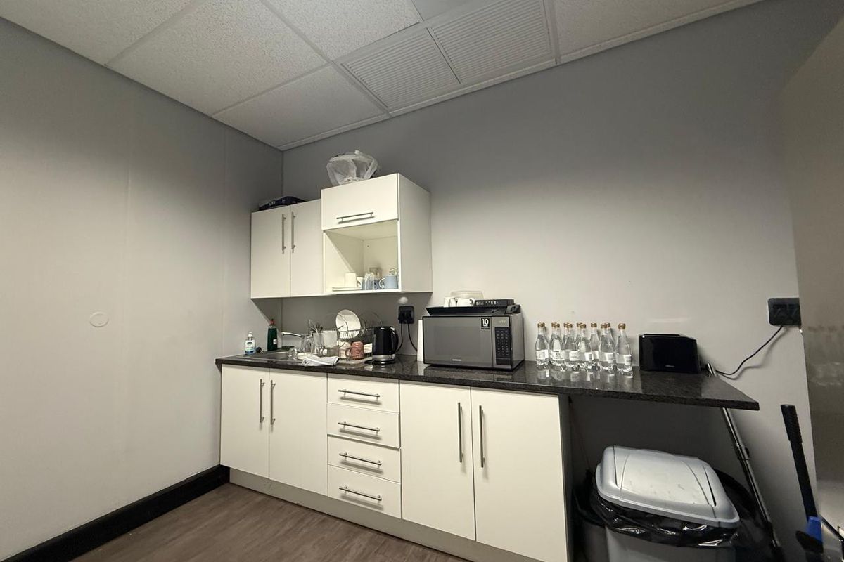 A functional office kitchenette featuring white cabinetry, a dark countertop, a stainless steel sink, and a drop ceiling with integrated lighting.