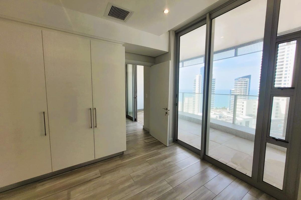 Modern bedroom featuring built-in cabinetry, wood-look tile flooring, and large sliding glass doors opening onto a private balcony with city and ocean views.