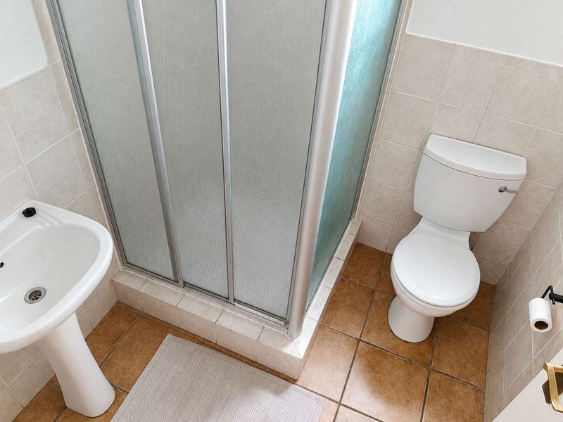 A functional bathroom featuring a pedestal basin, a corner shower enclosure, and a standard toilet with neutral wall and floor tiling.