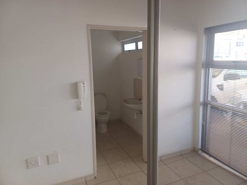 A functional industrial office bathroom featuring a toilet, wall-mounted basin, tiled flooring, and large windows with venetian blinds.