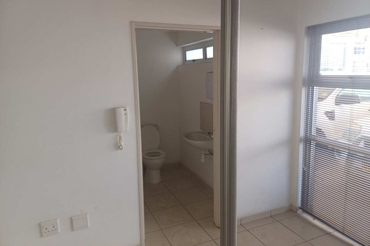 A functional industrial office bathroom featuring a toilet, wall-mounted basin, tiled flooring, and large windows with venetian blinds.