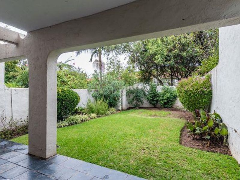 A covered patio area featuring tiled flooring and concrete pillars, overlooking a private garden with established landscaping and boundary walls.