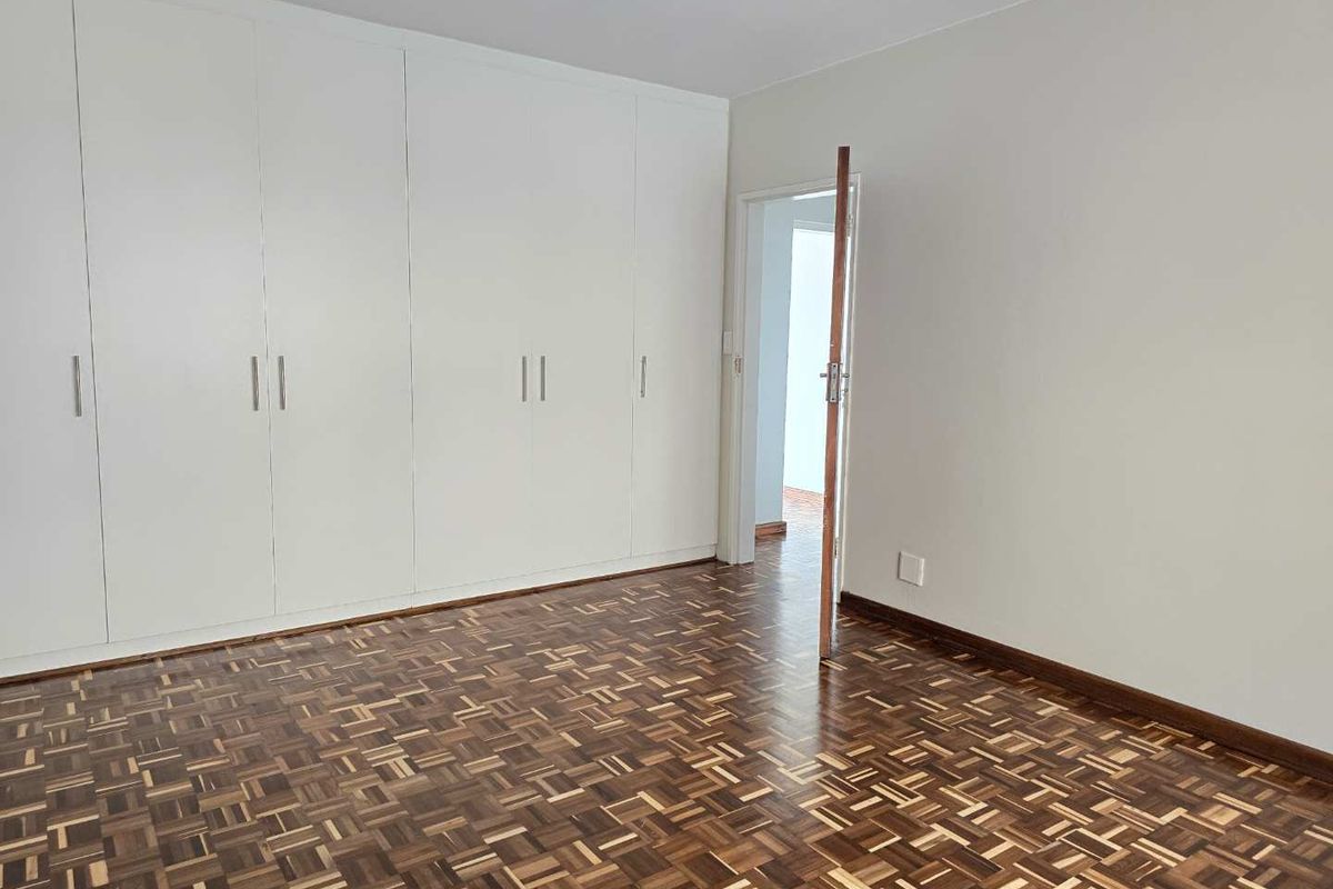 A bright, empty bedroom featuring well-maintained parquet flooring and extensive floor-to-ceiling built-in cupboards, offering ample storage space.