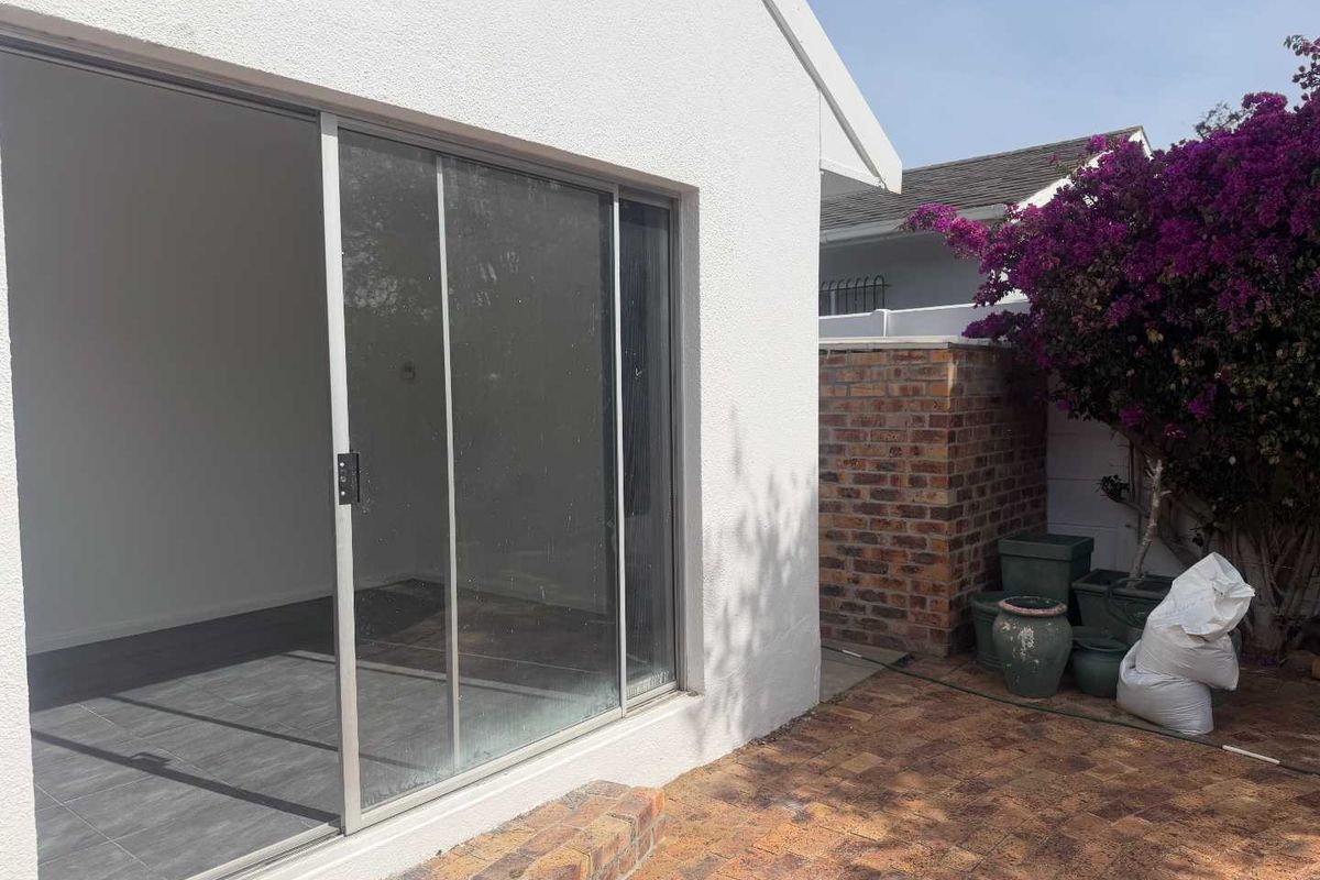 A rear exterior view featuring a paved patio area, sliding glass doors leading into the home, and a brick boundary wall with established bougainvillea.