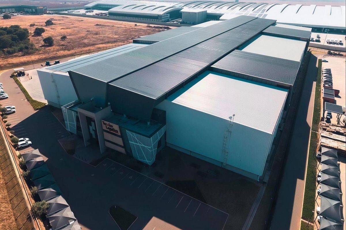 Aerial view of a modern industrial logistics facility featuring a large metal roof, extensive parking area, and secure perimeter fencing.