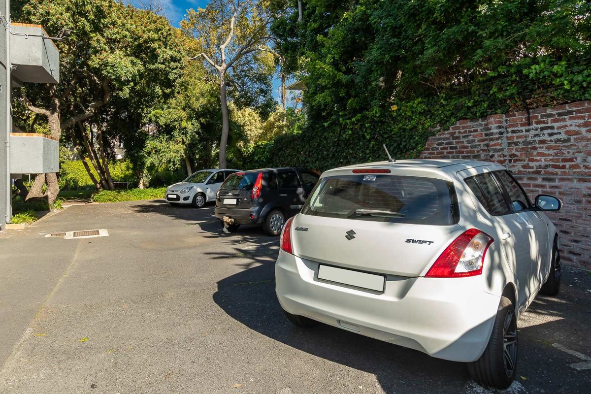 An outdoor parking area featuring an asphalt surface, brick boundary wall, and mature trees surrounding the property.