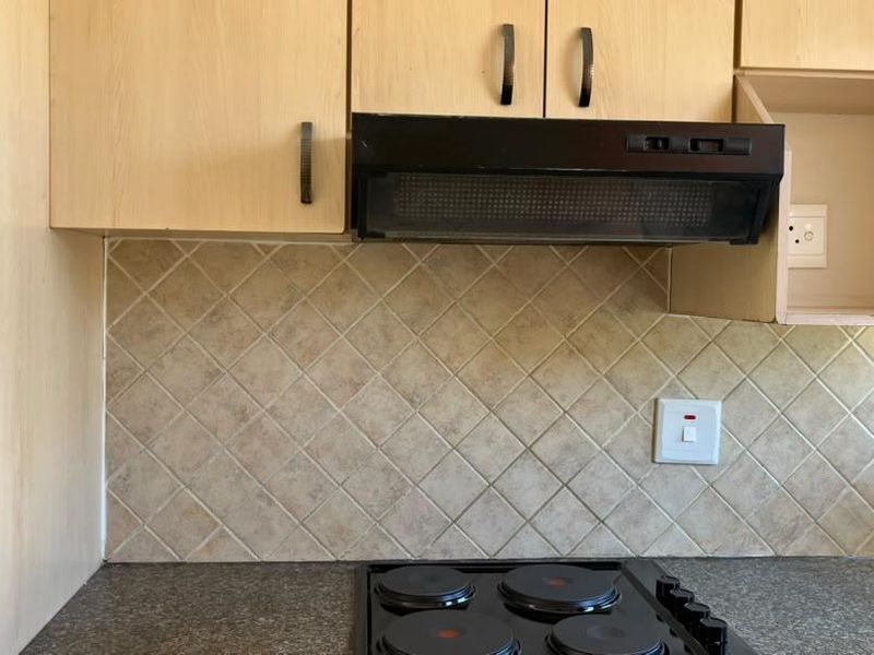 A functional kitchen area featuring light wood-finish cabinetry, a tiled backsplash, stone countertops, and a built-in electric hob with an extractor fan.