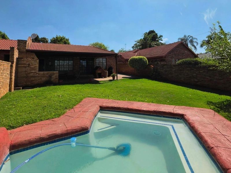 A private backyard featuring a swimming pool, a well-maintained lawn, and a covered patio area, enclosed by brick boundary walls.