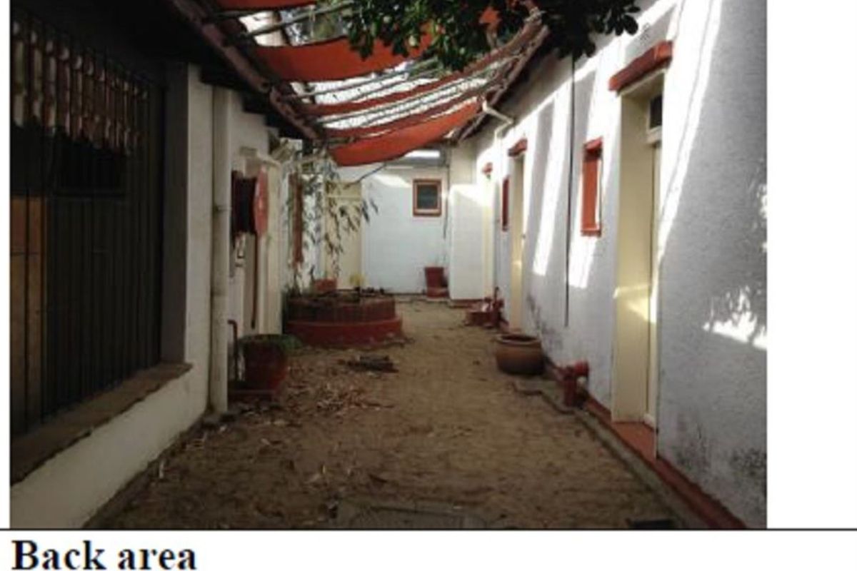 A narrow exterior rear courtyard area featuring a sand floor, multiple access doors, and a weathered overhead shade structure.