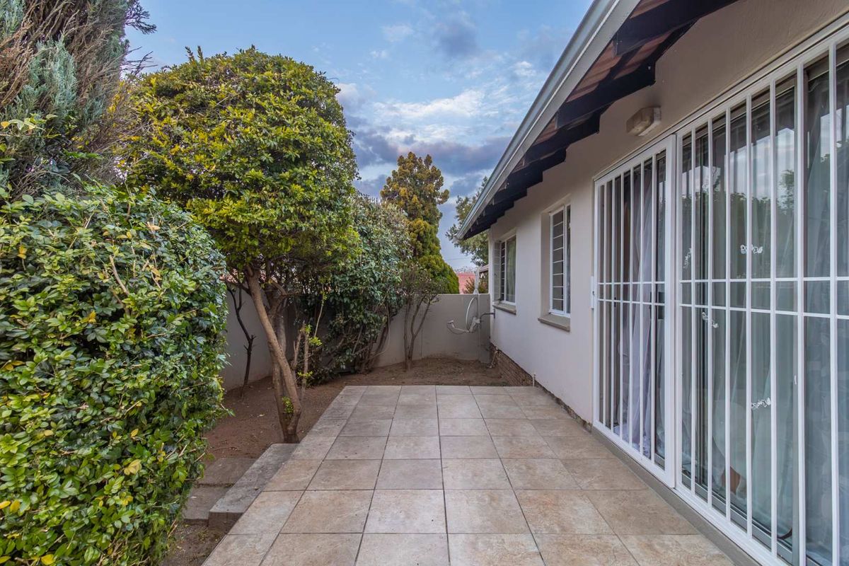 A paved rear patio area featuring sliding glass doors with security bars, overlooking a private garden space with established trees and boundary walls.