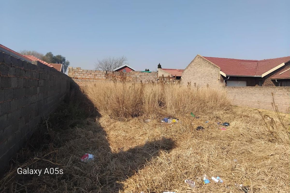 A vacant residential plot enclosed by perimeter masonry walls, featuring overgrown dry grass and accumulated debris.