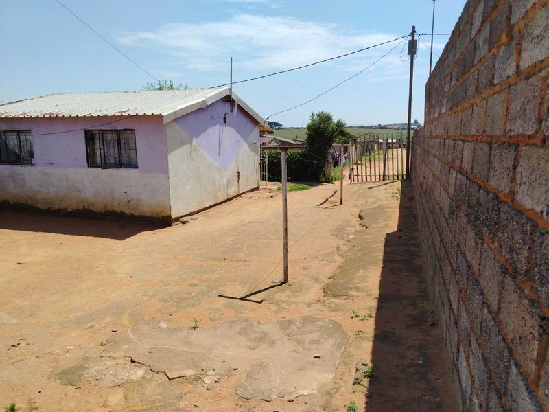 Rear exterior view of the property featuring a corrugated metal roof, perimeter brick wall, and an open yard area with concrete and dirt ground.