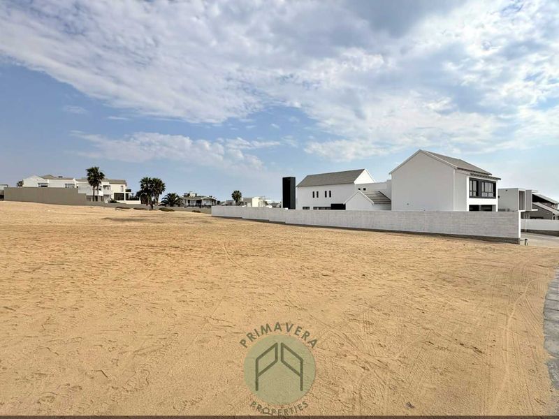 A vacant residential plot of land featuring a sandy surface, bordered by a boundary wall and paved street curb in a developed estate.