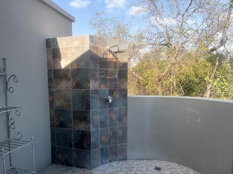 An outdoor shower area featuring a tiled enclosure, mosaic floor detailing, and a fixed shower head, set against a natural bushveld backdrop.