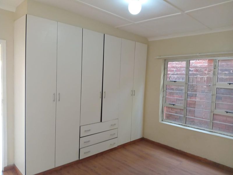 A clean, empty bedroom featuring white built-in cupboards, laminate flooring, and an aluminium window with security bars.