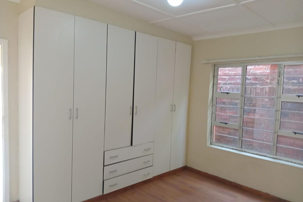 A clean, empty bedroom featuring white built-in cupboards, laminate flooring, and an aluminium window with security bars.