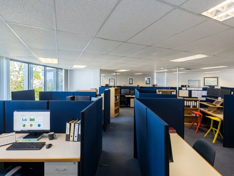 Open-plan office space featuring modular cubicle workstations, suspended ceilings with recessed lighting, and large curved windows providing natural light.