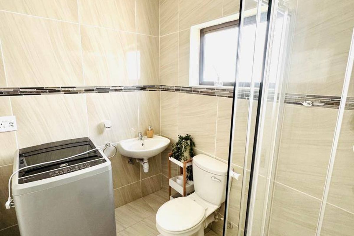 A functional bathroom featuring floor-to-ceiling wall tiles, a glass-enclosed shower, a wall-mounted basin, and a toilet, with dedicated space for laundry appliances.