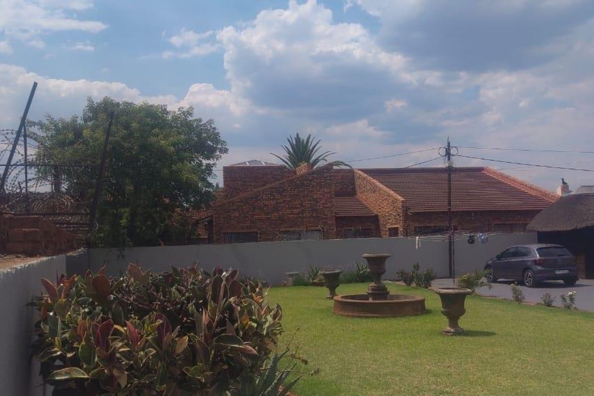 A well-maintained residential garden featuring a central stone fountain, manicured lawn, and perimeter boundary walls with security fencing.