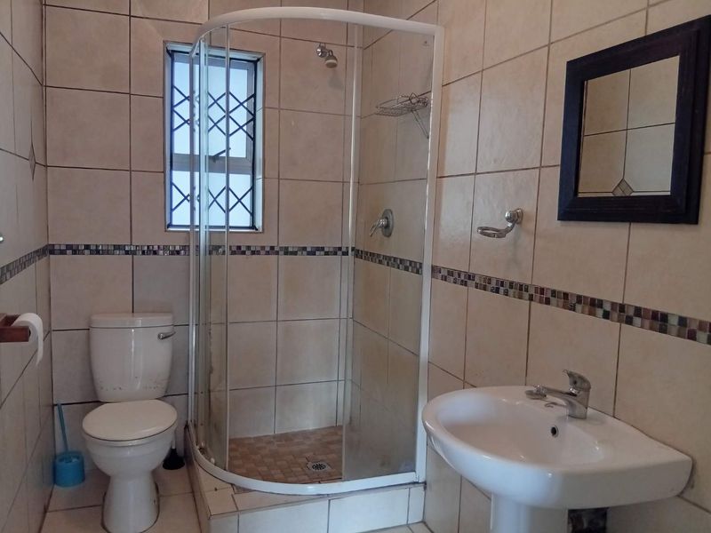 A functional bathroom featuring a corner shower enclosure, pedestal basin, and toilet with neutral wall tiling and security bars on the window.