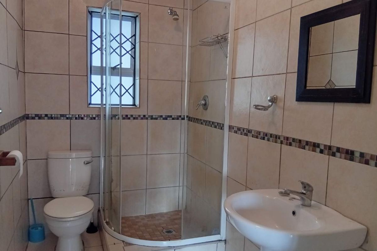 A functional bathroom featuring a corner shower enclosure, pedestal basin, and toilet with neutral wall tiling and security bars on the window.