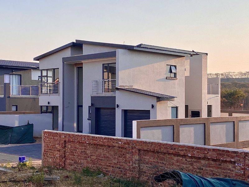 Modern double-story residential home featuring a double garage, balcony with metal railings, and a paved driveway, set within a secure perimeter wall.
