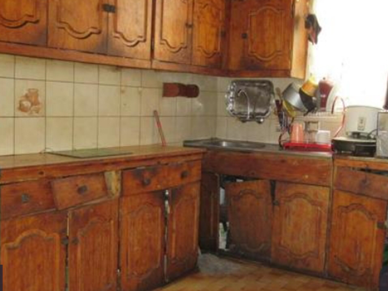 A dated kitchen featuring traditional wooden cabinetry, tiled backsplash, and a stainless steel sink, currently showing significant wear and tear on the fixed joinery.