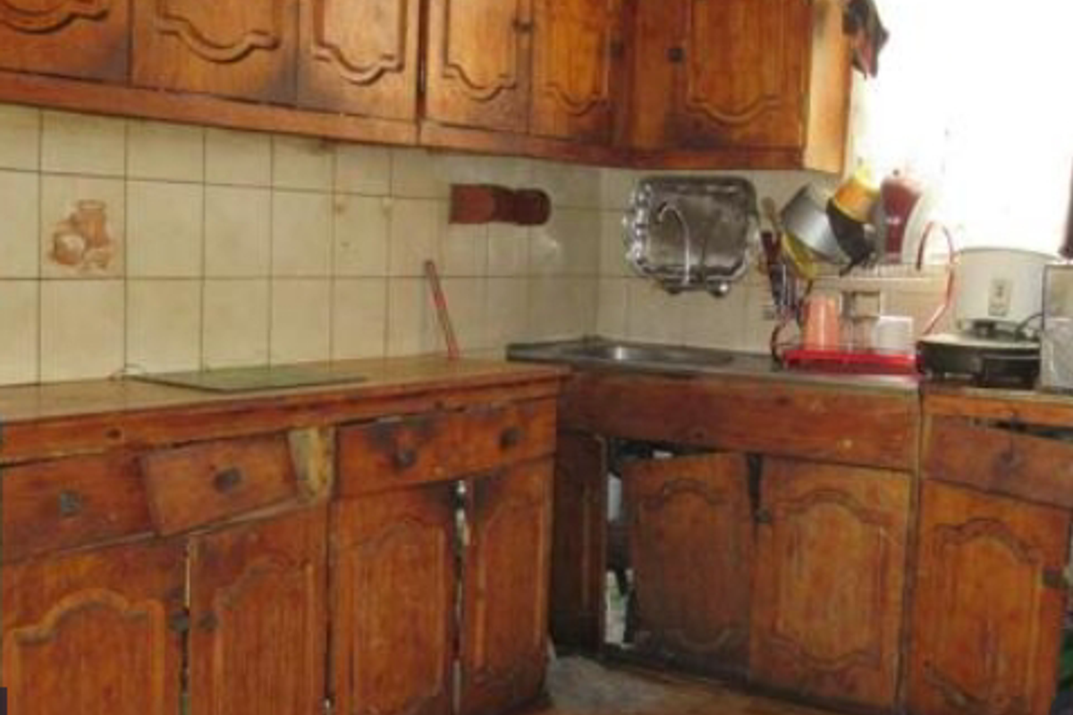 A dated kitchen featuring traditional wooden cabinetry, tiled backsplash, and a stainless steel sink, currently showing significant wear and tear on the fixed joinery.