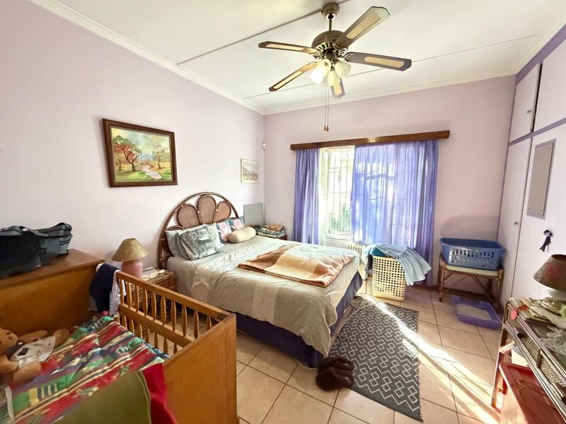 A bedroom featuring tiled flooring, a ceiling fan, and built-in cupboards, with natural light entering through a window fitted with security bars.