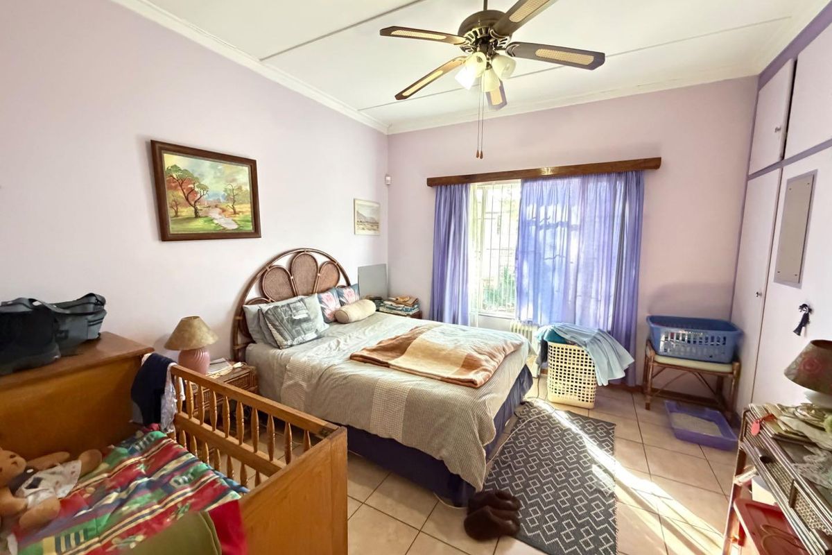 A bedroom featuring tiled flooring, a ceiling fan, and built-in cupboards, with natural light entering through a window fitted with security bars.