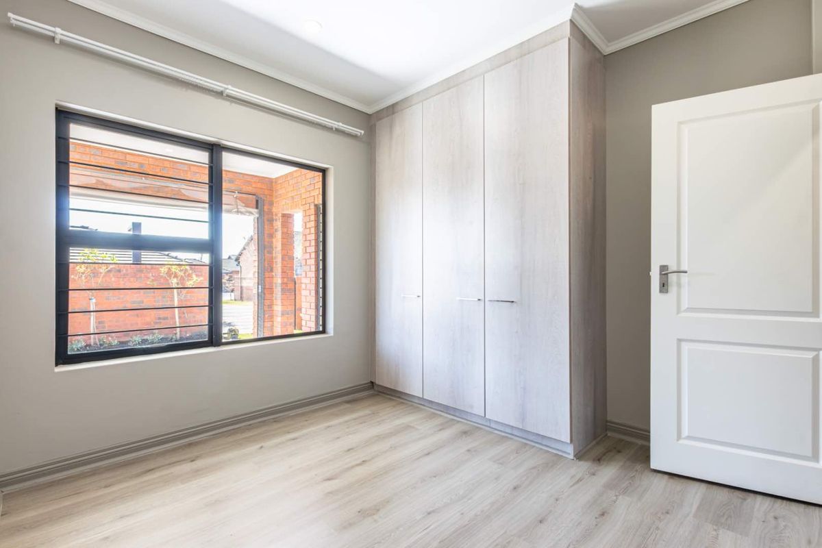 A bright, empty bedroom featuring modern light-toned laminate flooring, built-in wardrobes, and a large window with security bars.
