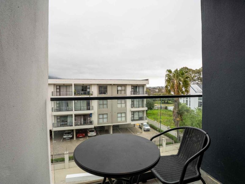 A private balcony featuring a modern glass balustrade with a metal handrail, offering an elevated view of the surrounding apartment complex and grounds.