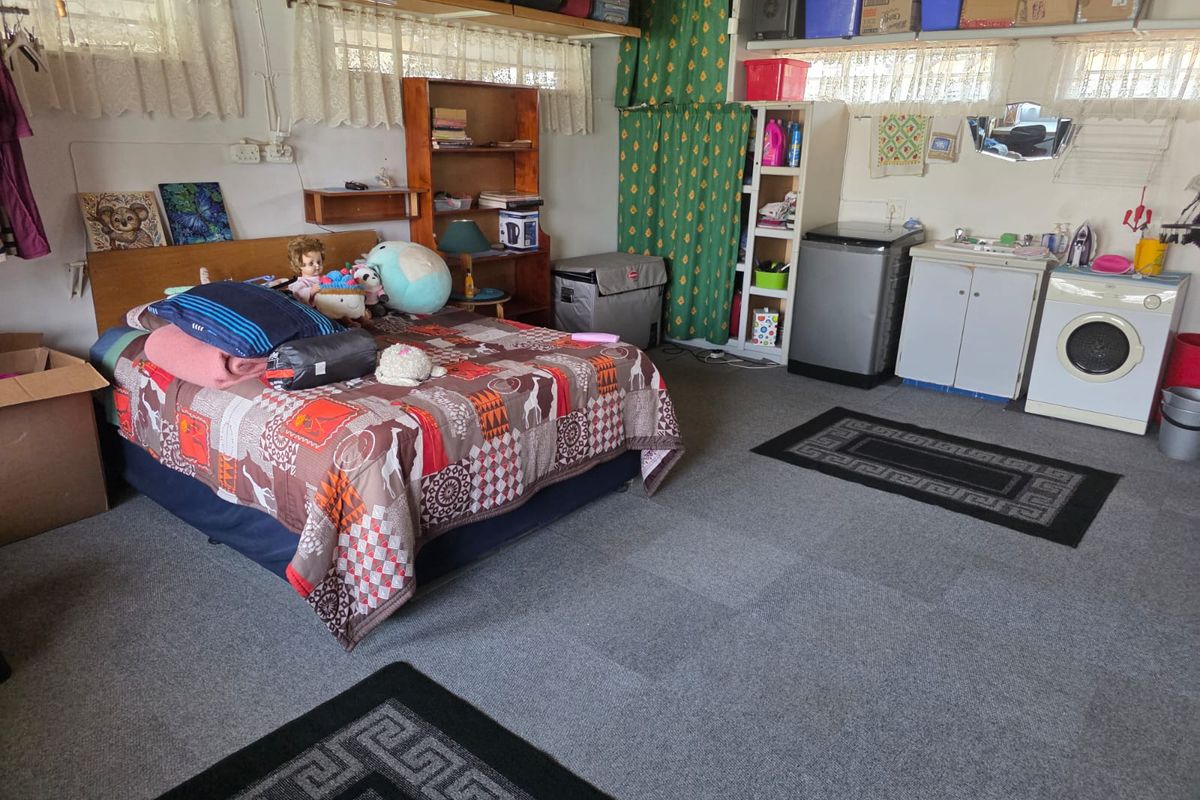 A multi-purpose bedroom space featuring integrated laundry facilities, including a sink and washing machine, with carpet flooring and overhead storage shelving.